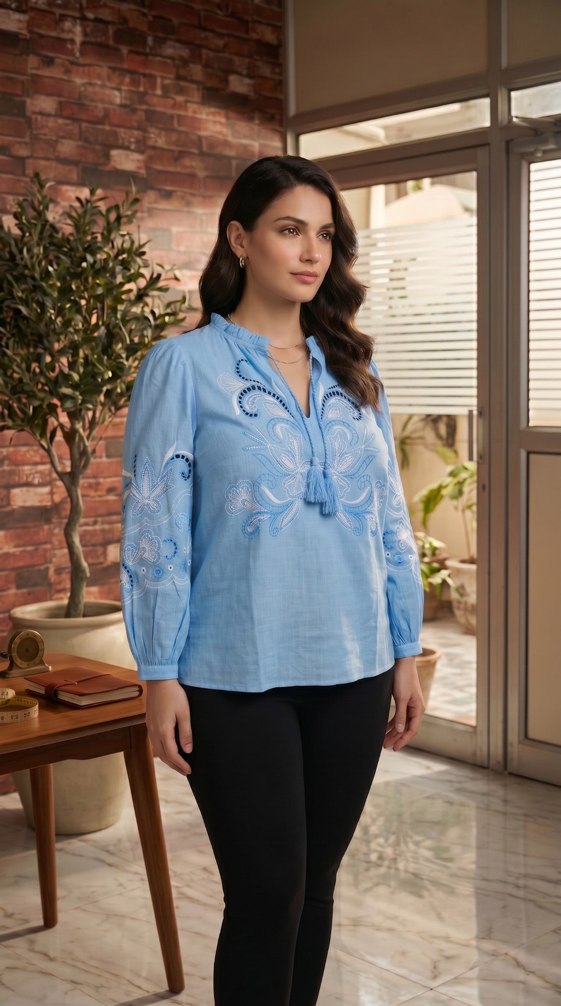 Woman wearing a sky blue embroidered top in an indoor setting with a plant and table in the background.